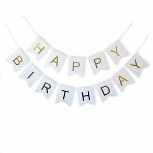 May include: A white banner with gold lettering that reads "HAPPY BIRTHDAY". The banner is made of small, flag-shaped pieces strung together with a thin white string. The letters are in a simple, sans-serif font.