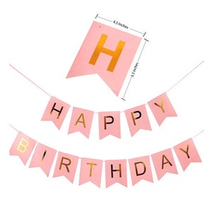 May include: Pink banner with gold lettering that reads "Happy Birthday." The banner is made of pink paper and has gold lettering. The banner is 6.5 inches long and 4.5 inches wide. The banner is for a birthday party.