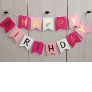 May include: A festive banner with the words "HAPPY BIRTHDAY" spelled out on colorful pennants. The pennants are in shades of pink, white, and gold, strung together with a white string. Perfect for birthday celebrations.