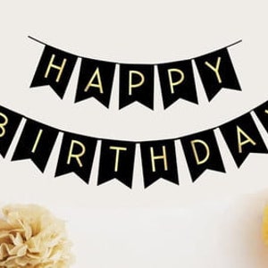 May include: Black banner flags spelling "HAPPY BIRTHDAY" in gold letters, strung across a white background. The flags are in a pennant shape. Yellow and beige paper flowers are in the foreground.