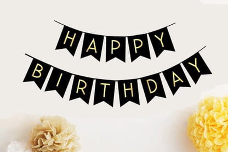 May include: Black banner flags spelling "HAPPY BIRTHDAY" in gold letters, strung across a white background. The flags are in a pennant shape. Yellow and beige paper flowers are in the foreground.
