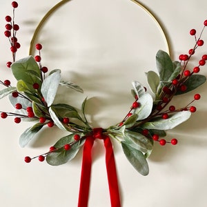 May include: A gold metal wreath with red berries and green leaves. The wreath has a red velvet ribbon attached to it. The text "May Home Studio" is written on the bottom of the image.