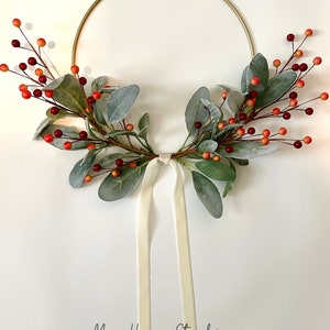 May include: A gold metal wreath with a white ribbon and faux greenery with red and orange berries.