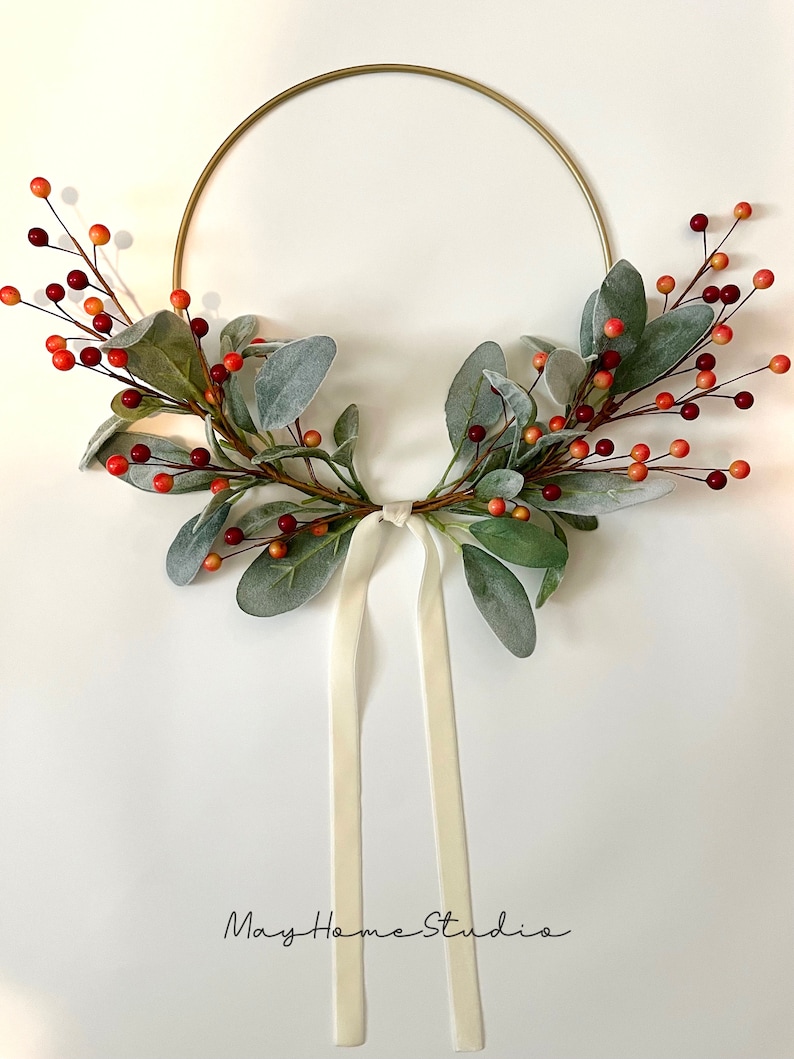 May include: A gold metal wreath with a white ribbon and faux greenery with red and orange berries.