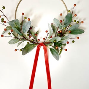 May include: A gold metal hoop wreath decorated with green and red faux greenery and berries. A red velvet ribbon is tied at the bottom of the wreath.