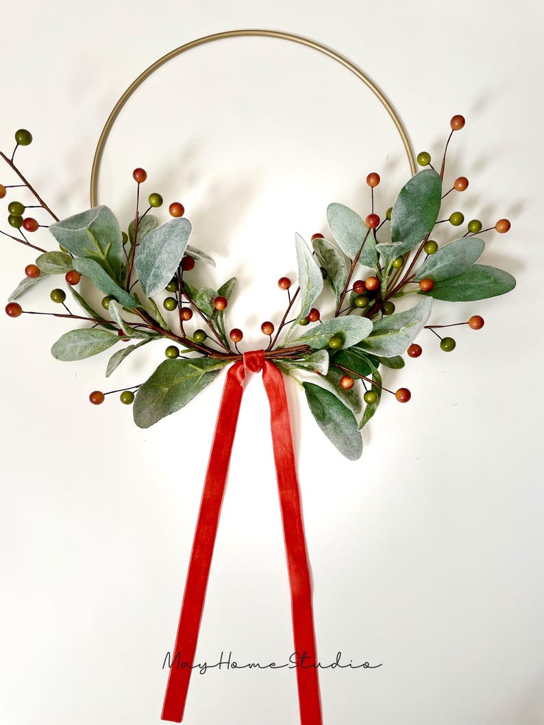 May include: A gold metal hoop wreath decorated with green and red faux greenery and berries. A red velvet ribbon is tied at the bottom of the wreath.