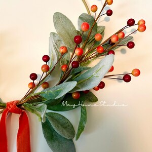 May include: A gold metal hoop wreath with a sprig of green leaves and red and orange berries. A red velvet ribbon is tied to the wreath.