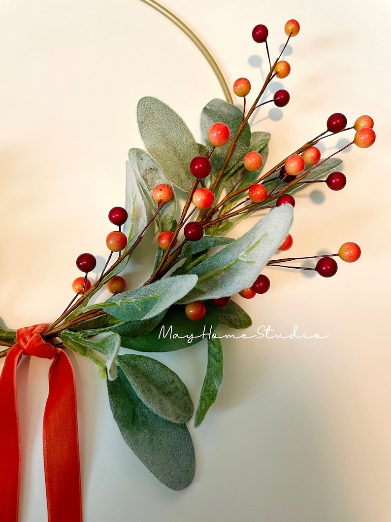 May include: A gold metal hoop wreath with a sprig of green leaves and red and orange berries. A red velvet ribbon is tied to the wreath.
