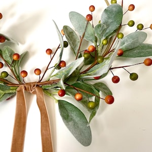 May include: A gold metal hoop wreath decorated with green and gray leaves and small red and orange berries. A brown velvet ribbon is tied at the bottom of the wreath.