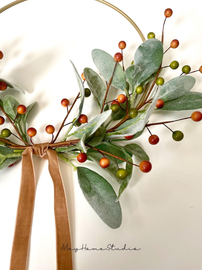 May include: A gold metal hoop wreath decorated with green and gray leaves and small red and orange berries. A brown velvet ribbon is tied at the bottom of the wreath.