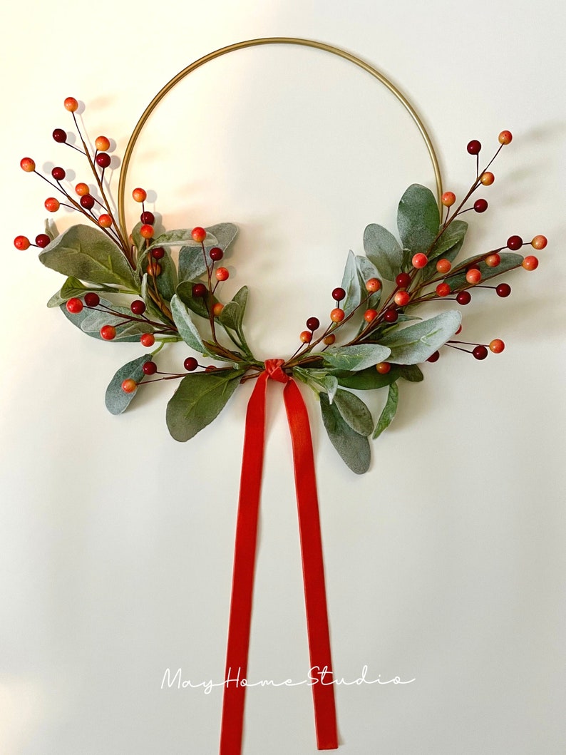 May include: A gold metal wreath with red and orange berries and green leaves. A red velvet ribbon hangs from the bottom of the wreath.