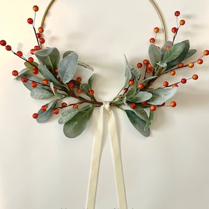 May include: A gold metal wreath with a white satin ribbon. The wreath is decorated with green leaves and red berries.
