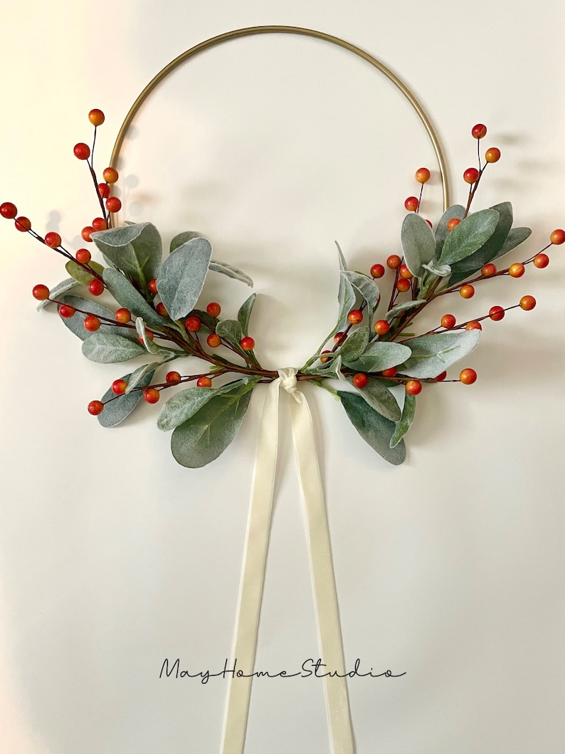 May include: A gold metal wreath with a white satin ribbon. The wreath is decorated with green leaves and red berries.