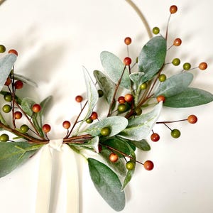 May include: A gold metal hoop wreath decorated with faux greenery and red berries. A white satin ribbon is tied at the bottom of the wreath.