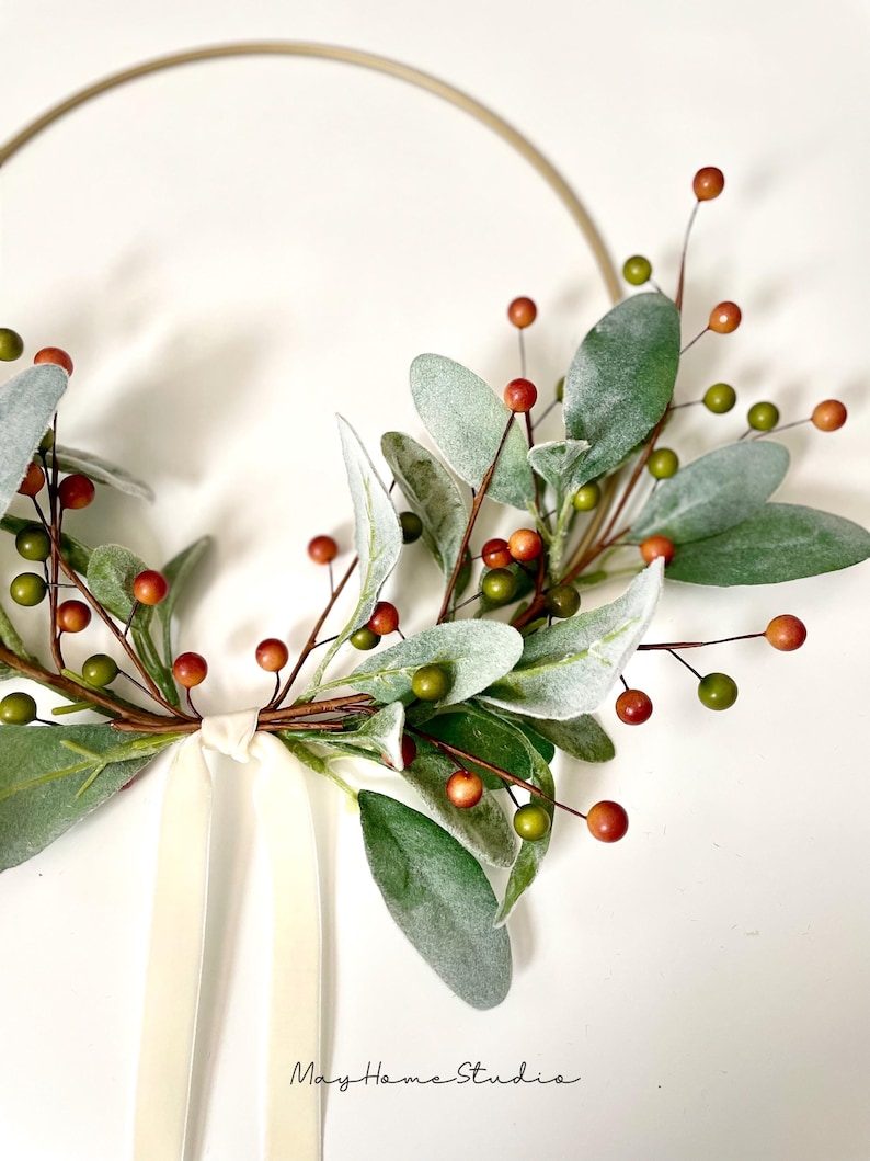 May include: A gold metal hoop wreath decorated with faux greenery and red berries. A white satin ribbon is tied at the bottom of the wreath.