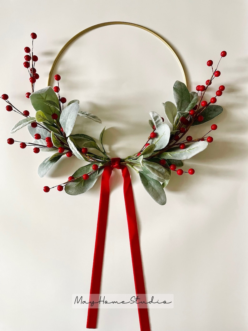 May include: A gold metal wreath with red berries and green leaves. The wreath has a red velvet ribbon attached to it. The text "May Home Studio" is written on the bottom of the image.