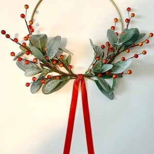 May include: A gold metal hoop wreath decorated with sprigs of faux greenery and red berries. A red velvet ribbon hangs from the bottom of the wreath.