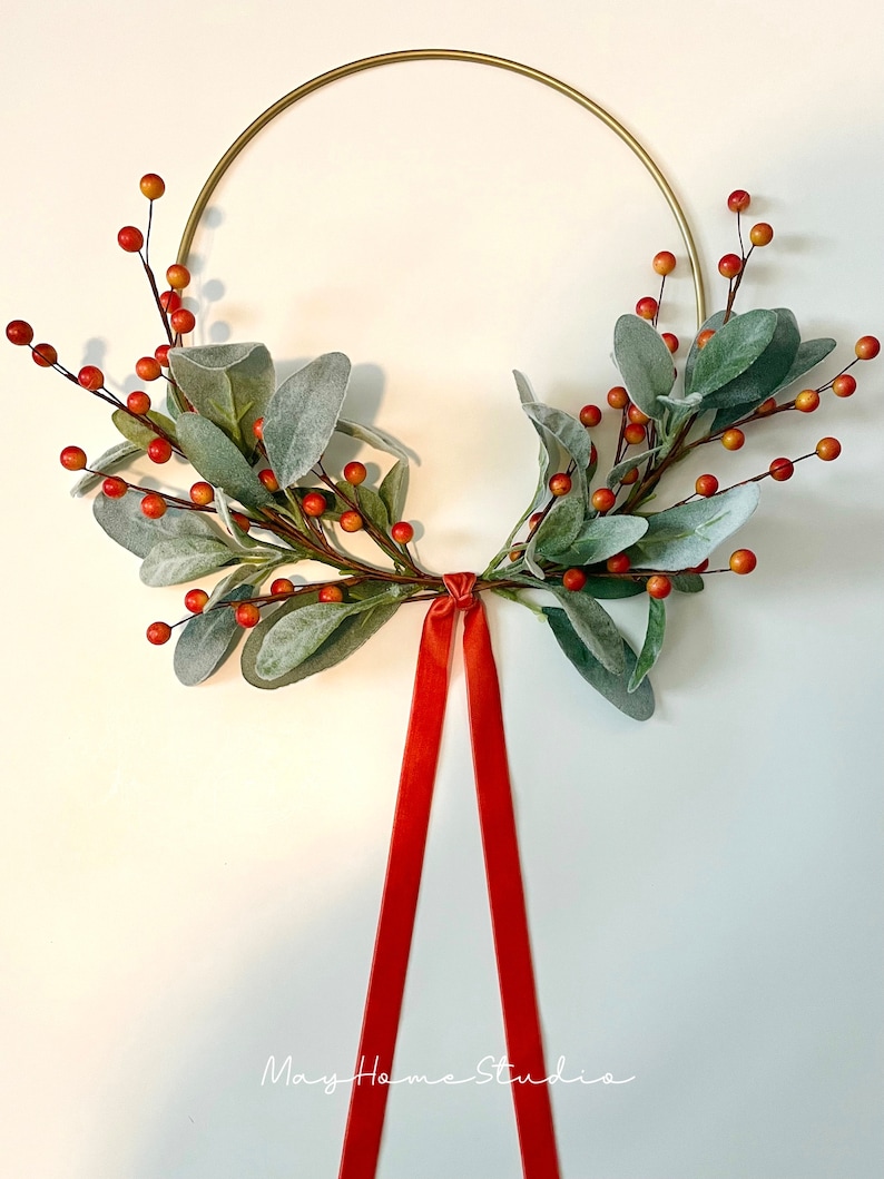 May include: A gold metal hoop wreath decorated with sprigs of faux greenery and red berries. A red velvet ribbon hangs from the bottom of the wreath.