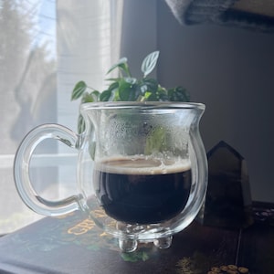 Pode incluir: Uma caneca de vidro transparente de parede dupla com al&ccedil;a, cheia de caf&eacute; escuro. A caneca est&aacute; sobre um livro com capa marrom escura e letras douradas.
