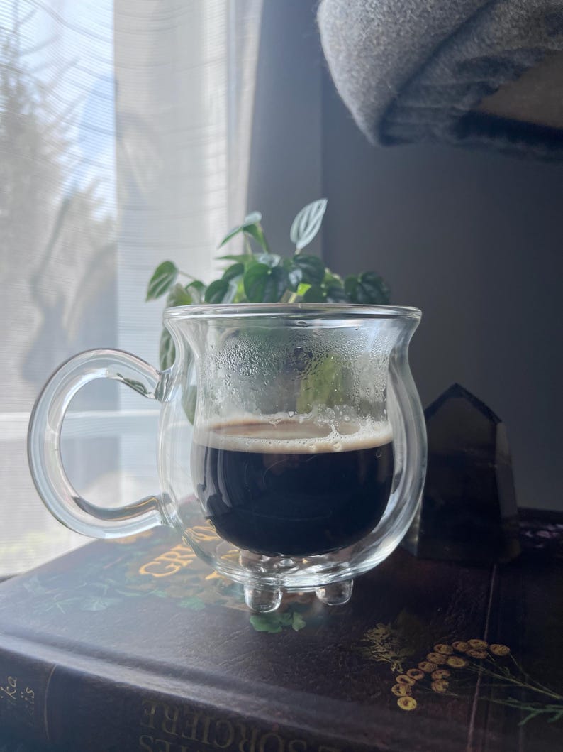 Pode incluir: Uma caneca de vidro transparente de parede dupla com al&ccedil;a, cheia de caf&eacute; escuro. A caneca est&aacute; sobre um livro com capa marrom escura e letras douradas.