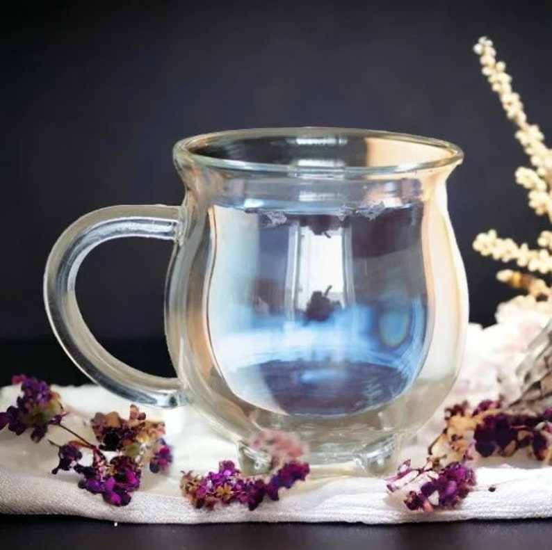 Pode incluir: Uma caneca de vidro transparente com corpo arredondado e al&ccedil;a. A caneca est&aacute; cheia de um l&iacute;quido azul claro. A caneca est&aacute; sobre uma superf&iacute;cie branca com flores roxas.