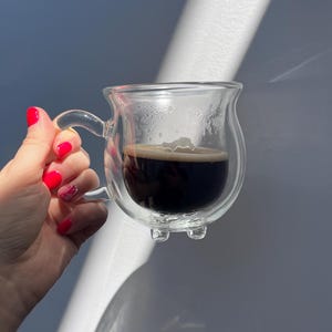 Pode incluir: Uma caneca de vidro transparente de parede dupla, cheia de caf&eacute; escuro. A caneca tem uma al&ccedil;a e um fundo arredondado com tr&ecirc;s pequenos p&eacute;s.