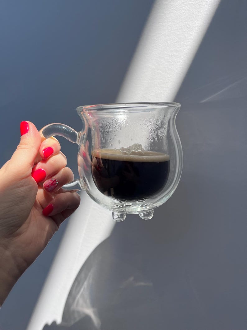 Pode incluir: Uma caneca de vidro transparente de parede dupla, cheia de caf&eacute; escuro. A caneca tem uma al&ccedil;a e um fundo arredondado com tr&ecirc;s pequenos p&eacute;s.