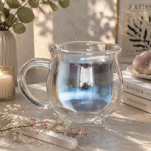 May include: A clear glass mug with a double-walled design and a rounded, cauldron-like shape. The mug features a curved handle and three small feet at the base. It is filled with a translucent blue liquid that reflects light, creating a soft glow.