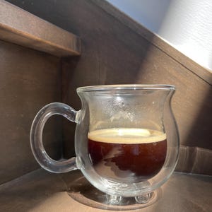 Pode incluir: Uma caneca de vidro transparente com parede dupla, cheia de caf&eacute; escuro. A caneca tem uma al&ccedil;a e um fundo arredondado.