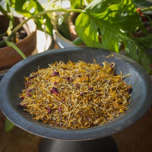 May include: A dark gray ceramic bowl filled with dried herbs and flower petals. The mixture is primarily yellow and orange, with small accents of purple and pink. The bowl sits on a dark pedestal, with green plants visible in the background.