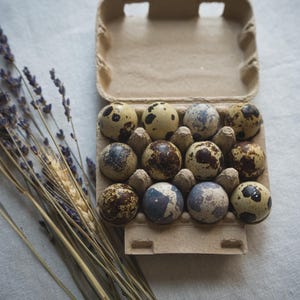 May include: A dozen speckled quail eggs in a cardboard egg carton. The eggs have brown, tan, and blue-grey markings. Dried lavender and wheat stalks are arranged to the left of the carton on a white linen surface.