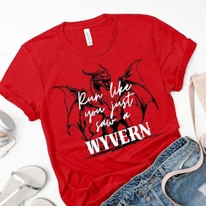 Pode incluir: Camiseta vermelha com um desenho de dragão preto e o texto branco "Run like you just saw a WYVERN". As mangas estão enroladas. A camiseta é apresentada com shorts jeans e acessórios.