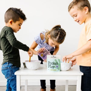 May include: Three children are making purple potion play with a packet of purple potion play powder. The packet has the text "Potion Play" and a purple handprint. The children are using wooden spoons to stir the powder into bowls.