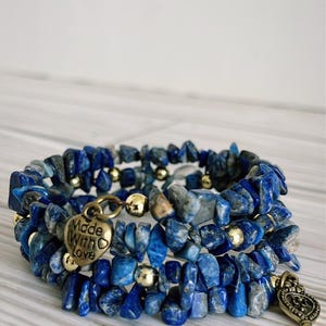 May include: A stack of three wrap bracelets made of blue lapis lazuli chips and gold-colored beads. Each bracelet has a charm, one with the words "Made With Love". The bracelets are on a light wood surface.