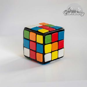 May include: A colourful Rubik's Cube cake, with red, blue, green, yellow, orange, and white squares. The cake is made of fondant and is shaped like a cube.