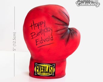 May include: A red boxing glove cake decorated with black writing that says "Happy Birthday Edvald" and a yellow Everlast logo. The cake is 5 inches tall (12.5 cm).
