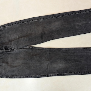May include: A pair of faded black denim jeans. The jeans feature a classic five-pocket design, a button fly, and a straight leg cut. The denim shows signs of wear and fading, giving them a vintage appearance. The jeans are laid flat on a light-coloured surface.
