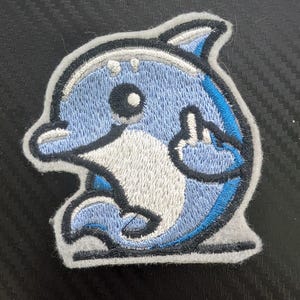 May include: Embroidered patch featuring a cartoon dolphin in shades of blue, white, and black. The dolphin has a friendly expression and is outlined in black with a gray border. The patch is likely for clothing or accessories.