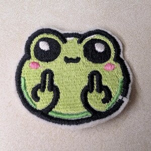 May include: Embroidered patch featuring a cartoon frog with a light green body, black eyes, and pink cheeks. The frog is giving the middle finger with both hands. The patch has a black border and a white backing.