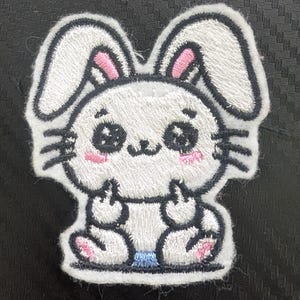 May include: Embroidered patch featuring a cartoon bunny with white fur, pink inner ears, and black outlines. The bunny has large, expressive eyes, pink cheeks, and is giving the middle finger with both hands. The patch has a white border.