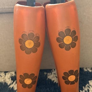 May include: Pair of orange boot shapers with black handles. Each shaper features three brown and yellow flower designs. The shapers are designed to maintain the shape of boots when stored.