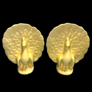 May include: A pair of pale yellow ceramic peacock figurines, each featuring a fanned tail with embossed feather details and a smooth, glossy finish. The birds are depicted in a standing position with wings tucked against their bodies.