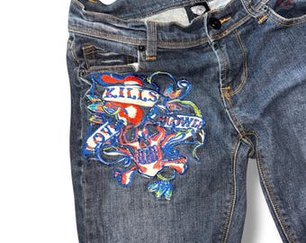 May include: Dark wash denim jeans featuring a prominent embroidered graphic on the front right thigh. The embroidery depicts a skull with blue and red accents and a banner with the text KILLS SLOWLY. The waistband includes a size 26 tag.