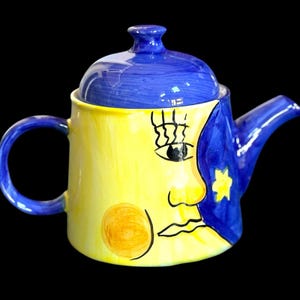 May include: A whimsical ceramic teapot with a blue lid and handle, and a spout. The teapot is painted with a yellow moon face design on one side and a blue night sky with a star on the other. The teapot has a round orange circle on the side.
