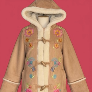 Op de afbeelding: Een bruine jas met een capuchon en knevelsluitingen. De jas heeft een crèmekleurige imitatiebontvoering en is versierd met kleurrijke geborduurde bloemenmotieven. De jas wordt gepresenteerd op een mannequin tegen een roze achtergrond.