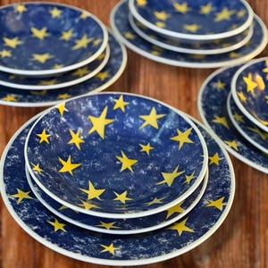May include: A set of blue ceramic plates featuring a yellow star design. The plates have a white rim and a distressed texture. The plates are stacked in sets of three, suitable for serving.