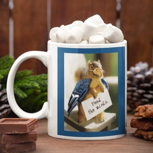 May include: White ceramic mug with a blue border around a picture of a squirrel wearing blue bird wings and holding a sign that says "Feed the Birds".
