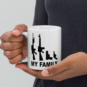 Pode incluir: Caneca de cerâmica branca com silhuetas pretas de armas de fogo e o texto "MY FAMILY". A caneca é segurada por uma pessoa. A caneca tem uma alça branca e é de tamanho padrão.