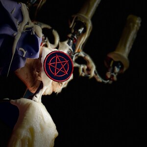 Può includere: Un candelabro con cera che cola e un'estetica oscura. Una toppa circolare con un pentagramma rosso su sfondo blu navy è attaccata al candelabro. Lo sfondo è nero.