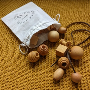 Pode incluir: Uma coleção de contas e formas de madeira natural, incluindo esferas, cubos e cilindros, espalhadas sobre uma superfície amarela texturizada. Uma bolsa branca com cordão com o texto "lachung designs" está parcialmente aberta, revelando mais peças de madeira. Um cordão de couro também está presente.